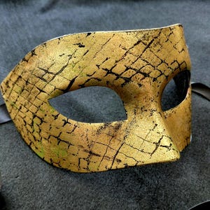 Op de afbeelding: Een goudkleurig masker voor een gemaskerd bal met een textuur, gebarsten patroon en zwarte accenten. Het masker heeft oogopeningen en is bevestigd aan een zwart lint. Het masker is ontworpen voor een kostuum of feest.