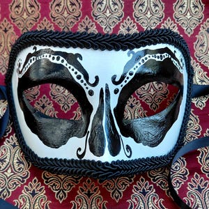 May include: A white masquerade mask featuring bold black painted designs around the eye sockets and nose area. The mask is edged with a thick black braided trim and includes black satin ribbons on both sides for securing the mask in place.
