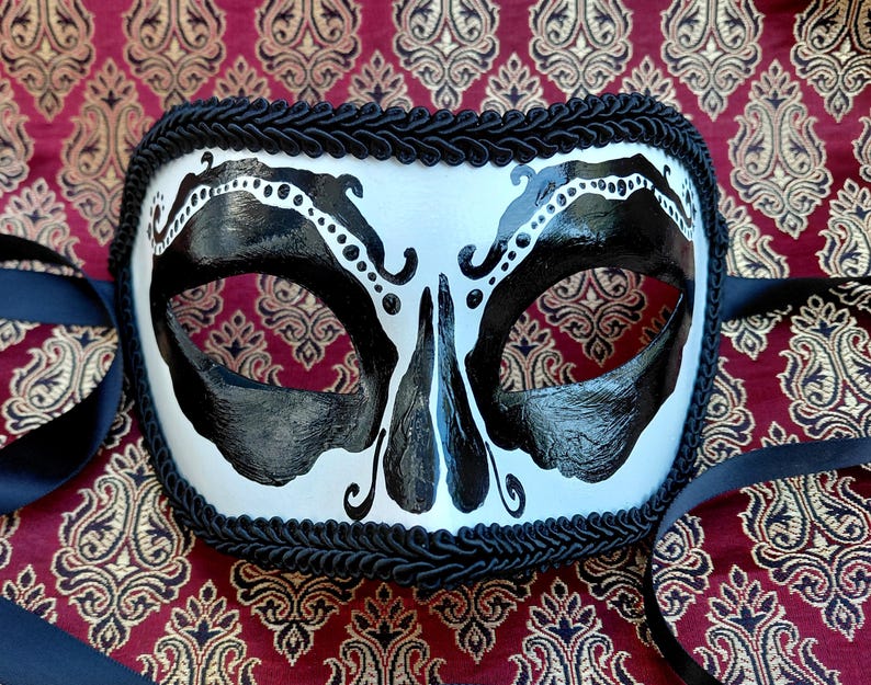 May include: A white masquerade mask featuring bold black painted designs around the eye sockets and nose area. The mask is edged with a thick black braided trim and includes black satin ribbons on both sides for securing the mask in place.