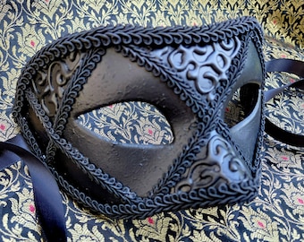 May include: A black masquerade mask featuring a textured surface with raised scrollwork patterns and intricate braided trim along the edges. The mask includes two eye openings and long black satin ribbons attached to the sides for securing it.