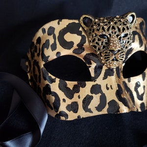 May include: A gold and black leopard print masquerade mask with a jeweled leopard head accent. The mask has black eye openings and black satin ribbons for tying. The mask is designed for a formal event or costume party.