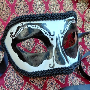 May include: A white masquerade mask featuring black painted accents around the eye openings and nose area, detailed with small white dots along the upper edges. The mask is bordered by a thick black braided trim and includes black ribbon ties.