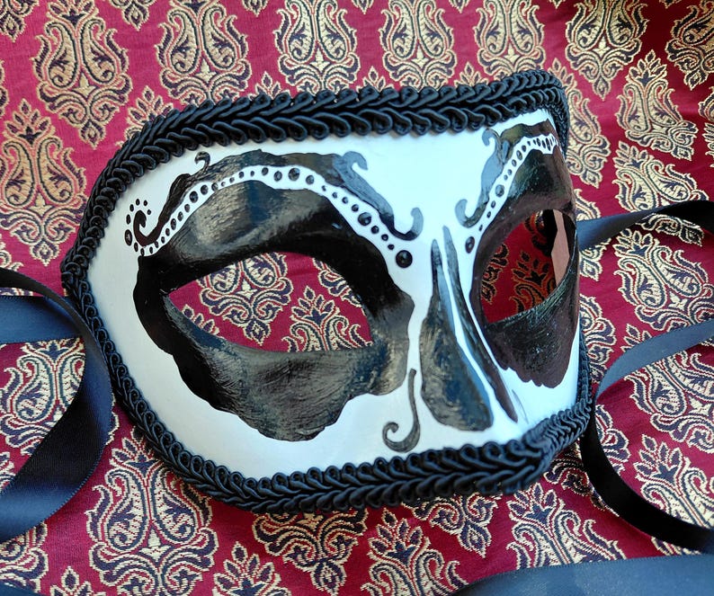 May include: A white masquerade mask featuring black painted accents around the eye openings and nose area, detailed with small white dots along the upper edges. The mask is bordered by a thick black braided trim and includes black ribbon ties.