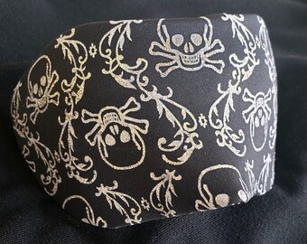 May include: Black fabric with a silver glitter skull and crossbones pattern. The fabric has a repeating pattern of skulls and crossbones with a floral design.