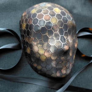 Op de afbeelding: Een decoratief masker met een honingraatpatroon in tinten zwart, bruin en goud. Het masker heeft een gladde, afgeronde vorm en is gemaakt van een metalen materiaal. Een zwart lint is om het masker gedrapeerd.