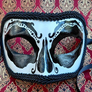 May include: A white masquerade mask featuring bold black painted accents around the eye openings and nose bridge. The mask is edged with a thick black braided trim and includes black satin ribbons on the sides for securing the piece to the face.