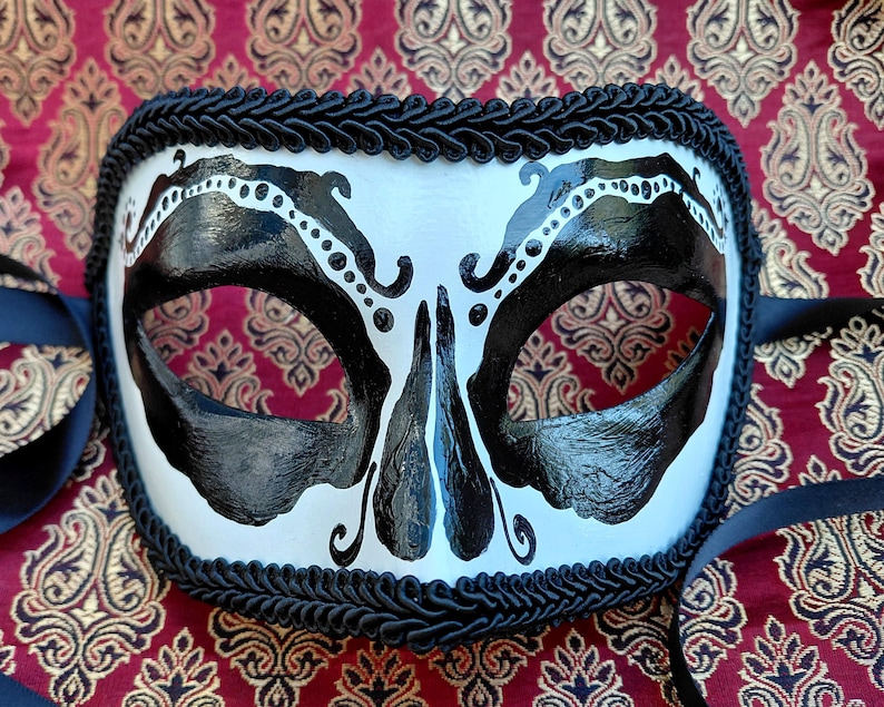 May include: A white masquerade mask featuring bold black painted accents around the eye openings and nose bridge. The mask is edged with a thick black braided trim and includes black satin ribbons on the sides for securing the piece to the face.