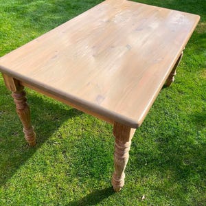 May include: A light brown wooden table with a rectangular top and turned legs. The table is set on grass. The table measures approximately 152 cm long and 76 cm wide.