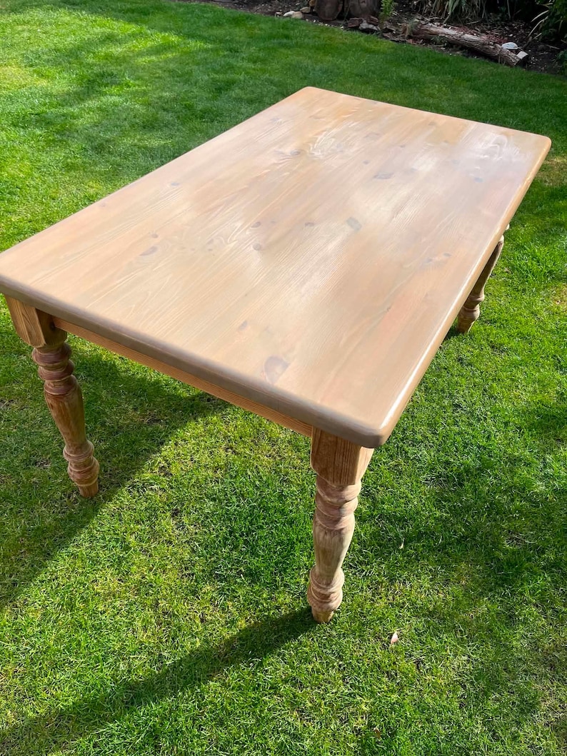 May include: A light brown wooden table with a rectangular top and turned legs. The table is set on grass. The table measures approximately 152 cm long and 76 cm wide.