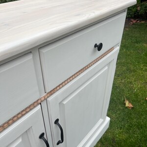 Puede incluir: Un mueble pintado de blanco con una tapa de madera clara. El mueble tiene dos puertas con tiradores negros y un caj&oacute;n con un pomo negro. Una moldura decorativa de madera recorre la parte delantera.