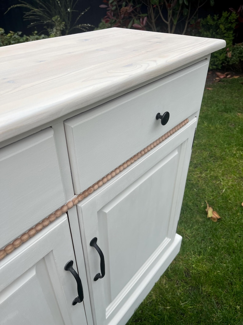 Puede incluir: Un mueble pintado de blanco con una tapa de madera clara. El mueble tiene dos puertas con tiradores negros y un caj&oacute;n con un pomo negro. Una moldura decorativa de madera recorre la parte delantera.
