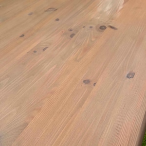 May include: Close-up of a light brown wooden table surface. The wood grain is visible, with knots and variations in colour. The table appears to be unfinished or lightly stained, showcasing the natural wood tones. The surface is smooth and appears to be recently constructed.