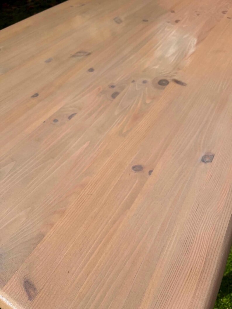 May include: Close-up of a light brown wooden table surface. The wood grain is visible, with knots and variations in colour. The table appears to be unfinished or lightly stained, showcasing the natural wood tones. The surface is smooth and appears to be recently constructed.