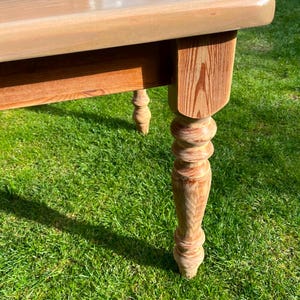 May include: A light-coloured wooden table with a rectangular top and turned legs. The table has a natural wood grain finish and is set on a green grassy surface. The legs are intricately carved.