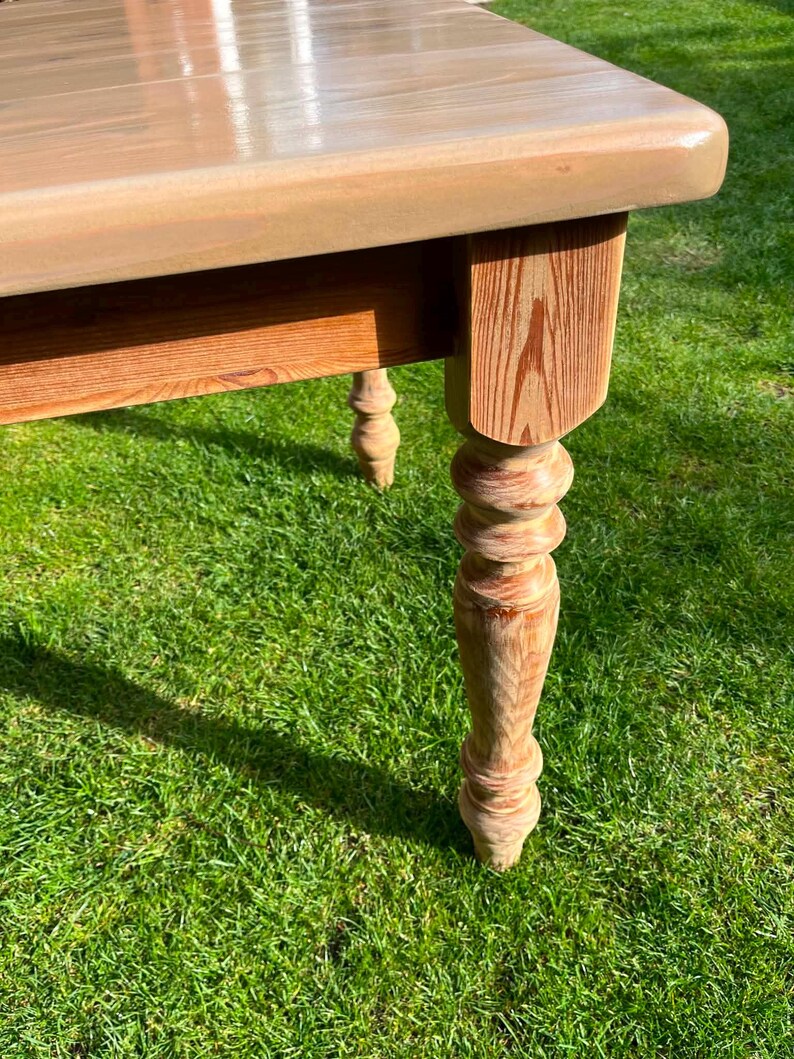 May include: A light-coloured wooden table with a rectangular top and turned legs. The table has a natural wood grain finish and is set on a green grassy surface. The legs are intricately carved.