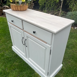 Puede incluir: Un mueble pintado de blanco con dos cajones y dos puertas, con herrajes negros. Una cesta tejida llena de flores coloridas se encuentra encima. El mueble est&aacute; sobre un c&eacute;sped verde.