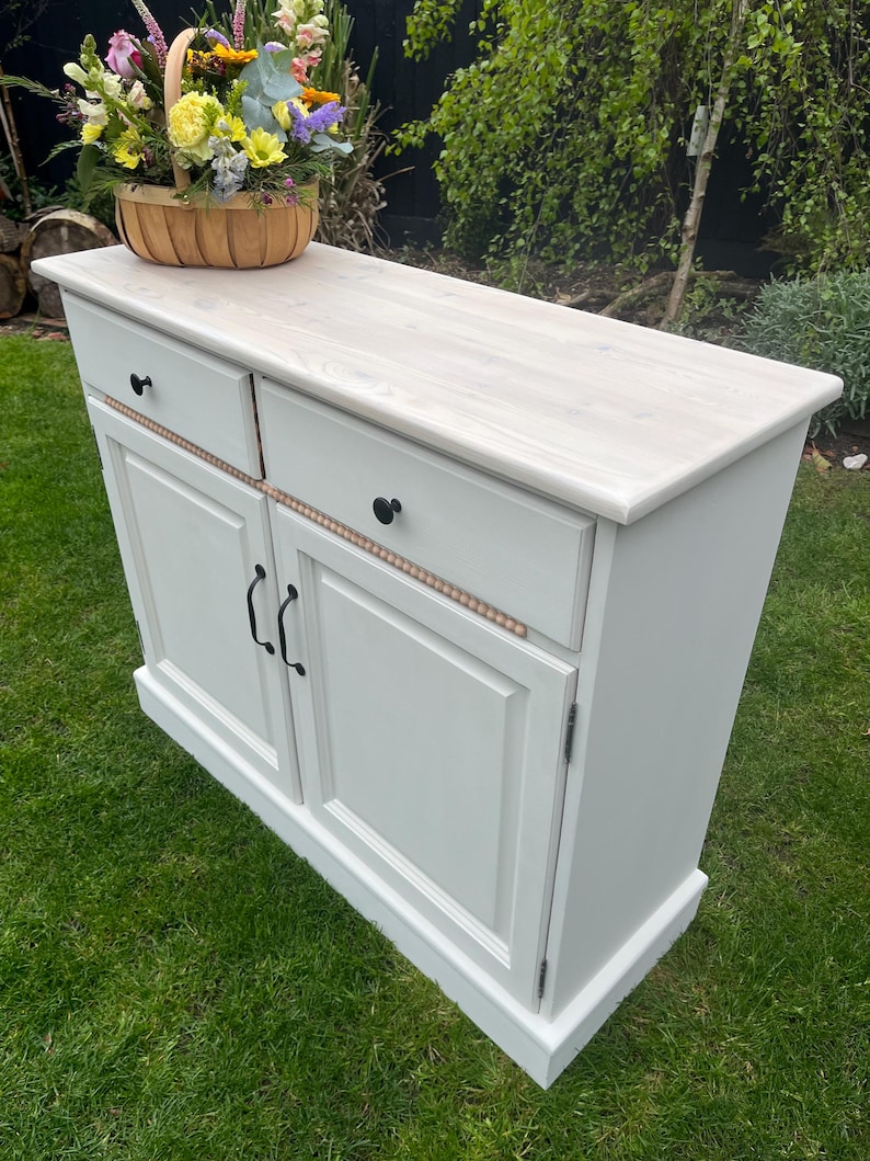 Puede incluir: Un mueble pintado de blanco con dos cajones y dos puertas, con herrajes negros. Una cesta tejida llena de flores coloridas se encuentra encima. El mueble est&aacute; sobre un c&eacute;sped verde.