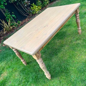 May include: A light-coloured wooden table with a rectangular top and turned legs. The table is set on a green lawn, highlighting its design. The table's dimensions are not visible, but it is a substantial piece of furniture. The table is made of wood.