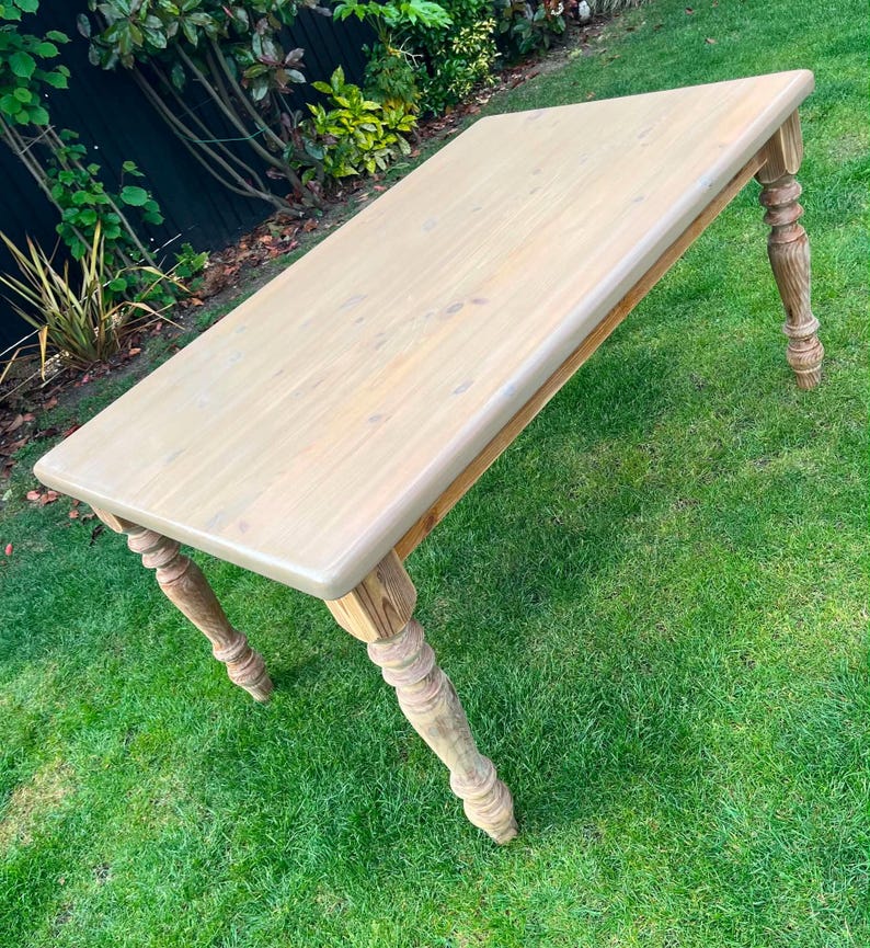 May include: A light-coloured wooden table with a rectangular top and turned legs. The table is set on a green lawn, highlighting its design. The table's dimensions are not visible, but it is a substantial piece of furniture. The table is made of wood.
