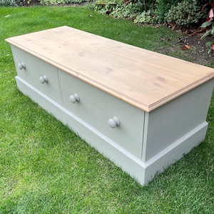 May include: A rectangular wooden storage chest featuring a natural light brown wood grain top surface and a light grey painted base. The front panel includes four small, round, light grey wooden knobs arranged in two pairs across the front.