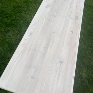 Puede incluir: Una mesa rectangular de madera clara con un acabado blanqueado. La veta de la madera es visible y la mesa est&aacute; sobre un fondo de hierba verde. La superficie de la mesa parece lisa y limpia.