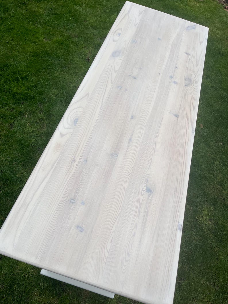 Puede incluir: Una mesa rectangular de madera clara con un acabado blanqueado. La veta de la madera es visible y la mesa est&aacute; sobre un fondo de hierba verde. La superficie de la mesa parece lisa y limpia.