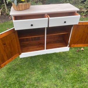 Puede incluir: Un aparador blanco con dos cajones y dos puertas. Las puertas y los cajones tienen pomos negros. El interior del mueble es de color marr&oacute;n c&aacute;lido, contrastando con el exterior blanco. Una cesta de flores est&aacute; encima.