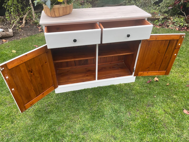 Puede incluir: Un aparador blanco con dos cajones y dos puertas. Las puertas y los cajones tienen pomos negros. El interior del mueble es de color marr&oacute;n c&aacute;lido, contrastando con el exterior blanco. Una cesta de flores est&aacute; encima.