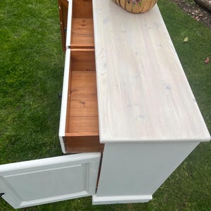 Puede incluir: Un mueble de madera pintado de blanco con dos cajones abiertos y una puerta. El mueble tiene una tapa de madera clara y una cesta de flores encima. Los cajones y el interior del mueble son de madera natural.