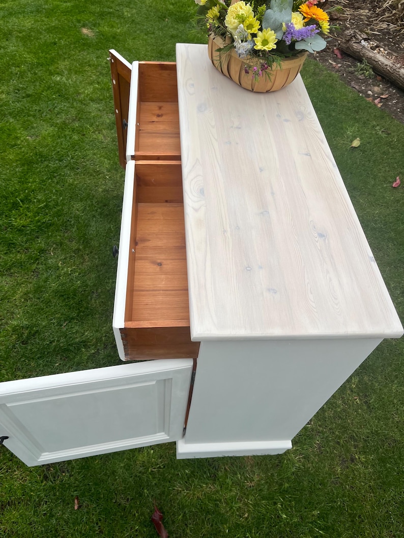 Puede incluir: Un mueble de madera pintado de blanco con dos cajones abiertos y una puerta. El mueble tiene una tapa de madera clara y una cesta de flores encima. Los cajones y el interior del mueble son de madera natural.