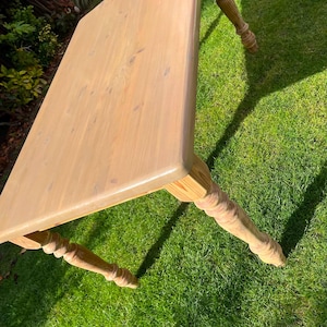May include: A light-coloured wooden table with a rectangular top and four turned legs. The table is made of unfinished wood and sits on a green grassy surface. The table's dimensions are approximately 122 cm long, 76 cm wide, and 76 cm high.