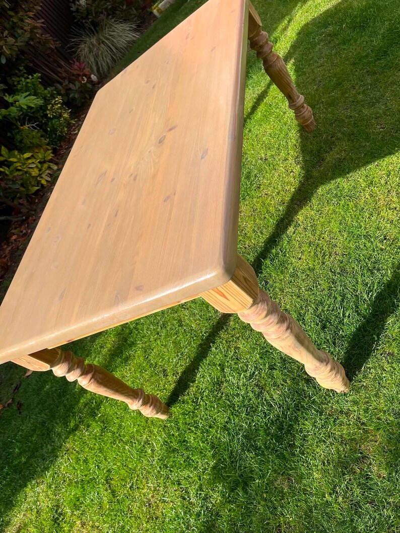 May include: A light-coloured wooden table with a rectangular top and four turned legs. The table is made of unfinished wood and sits on a green grassy surface. The table's dimensions are approximately 122 cm long, 76 cm wide, and 76 cm high.
