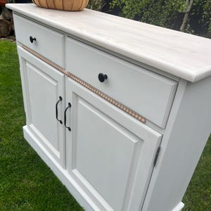 Puede incluir: Un aparador gris claro con dos cajones y dos puertas. El mueble tiene pomos y tiradores negros, y un ribete decorativo de cuentas. Una cesta de flores se encuentra encima del aparador.