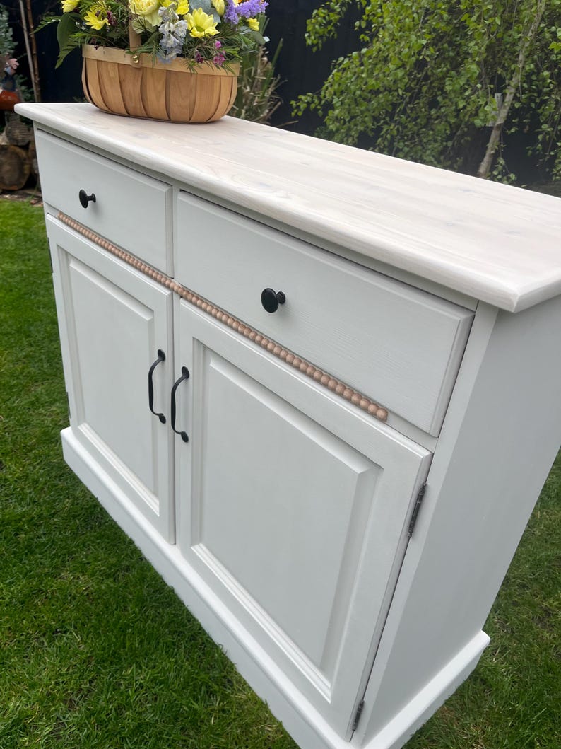 Puede incluir: Un aparador gris claro con dos cajones y dos puertas. El mueble tiene pomos y tiradores negros, y un ribete decorativo de cuentas. Una cesta de flores se encuentra encima del aparador.