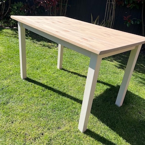 May include: A rectangular wooden table with a light-coloured tabletop and white legs. The table is set on a green lawn, with the sun casting shadows. The table's dimensions are approximately 152 cm long and 76 cm wide.