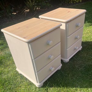 May include: Two bedside tables featuring a light-coloured natural wood top and a cream-coloured base. Each table has three drawers, each equipped with a single round knob. The units are rectangular in shape and stand on small, rounded feet.