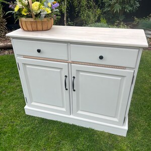 Puede incluir: Un aparador blanco con dos cajones y dos puertas, adornado con pomos y tiradores negros. Una moldura decorativa de cuentas de madera adorna los cajones. Una cesta de mimbre llena de flores coloridas se encuentra encima del aparador.