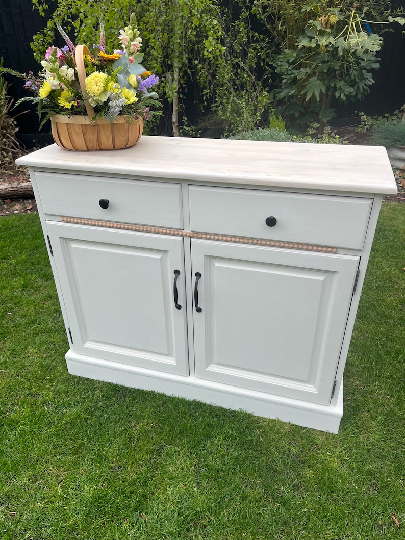 Puede incluir: Un aparador blanco con dos cajones y dos puertas, adornado con pomos y tiradores negros. Una moldura decorativa de cuentas de madera adorna los cajones. Una cesta de mimbre llena de flores coloridas se encuentra encima del aparador.