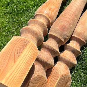 May include: A collection of unfinished wooden balusters, light brown in colour, resting on green grass. The balusters feature a turned design with a square base and decorative top, displaying the wood grain.