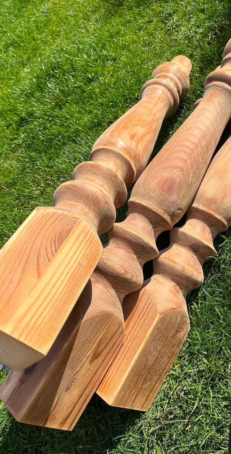 May include: A collection of unfinished wooden balusters, light brown in colour, resting on green grass. The balusters feature a turned design with a square base and decorative top, displaying the wood grain.