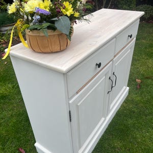 Puede incluir: Un mueble blanco con una encimera de madera clara y herrajes negros. Una cesta tejida llena de flores coloridas encima. El mueble tiene dos cajones y dos puertas.