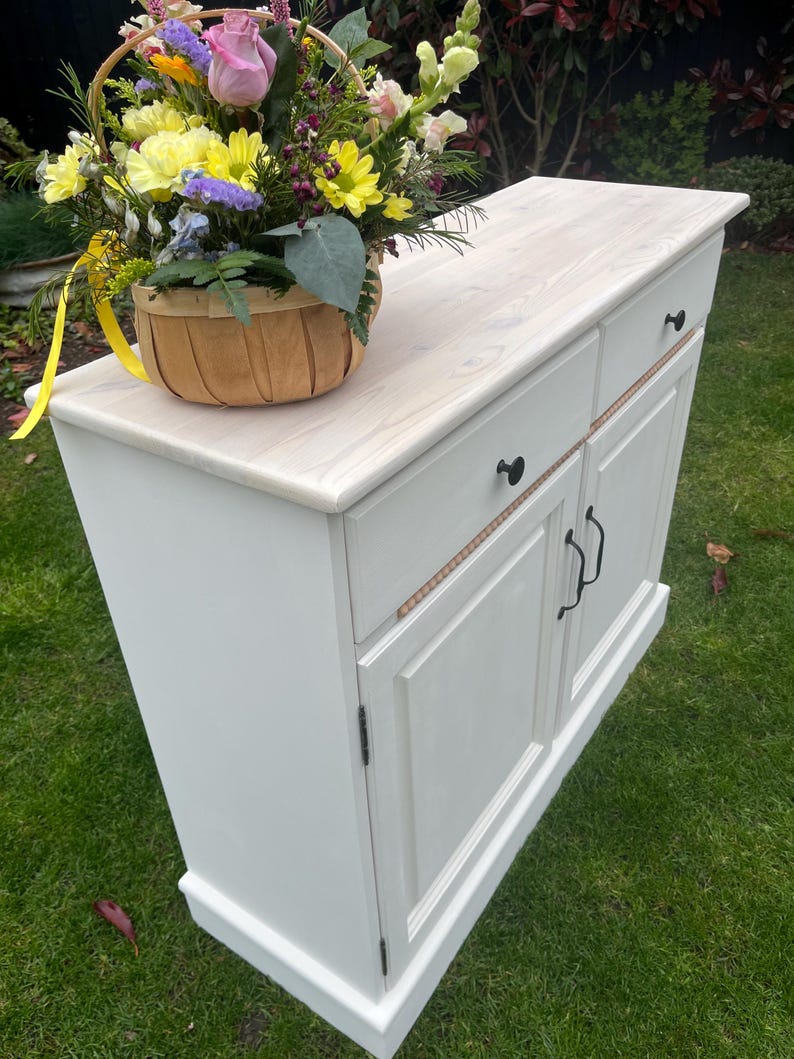 Puede incluir: Un mueble blanco con una encimera de madera clara y herrajes negros. Una cesta tejida llena de flores coloridas encima. El mueble tiene dos cajones y dos puertas.