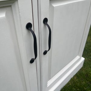 Puede incluir: Primer plano de un armario blanco con dos puertas y tiradores negros. Las puertas del armario tienen un dise&ntilde;o de panel en relieve. Los tiradores negros son curvos y tienen un extremo decorativo y redondeado. El armario est&aacute; sobre hierba verde.