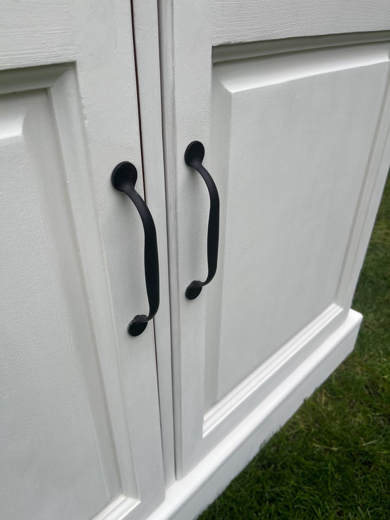 Puede incluir: Primer plano de un armario blanco con dos puertas y tiradores negros. Las puertas del armario tienen un dise&ntilde;o de panel en relieve. Los tiradores negros son curvos y tienen un extremo decorativo y redondeado. El armario est&aacute; sobre hierba verde.