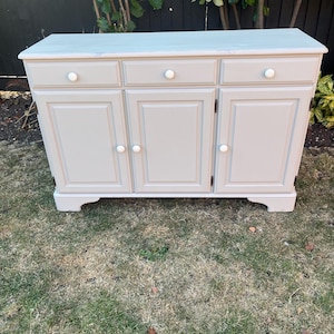 May include: A light grey sideboard featuring three top drawers and three lower cabinet doors. Each drawer and door is equipped with a small, round white knob. The piece has a rectangular top surface and a base with cutout feet at the corners.