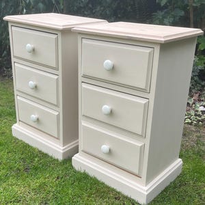 May include: A pair of matching bedside cabinets featuring three drawers each. The units are painted in a light beige finish with a natural wood top surface. Each drawer is equipped with a small, round, white knob for opening and closing the drawers.