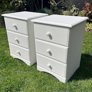 May include: A pair of white bedside cabinets, each with three drawers and round knobs. The cabinets are positioned on grass, displaying a traditional design. Each drawer has a recessed panel.