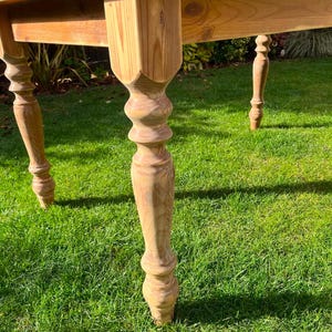 May include: A light brown wooden table with a rectangular top and four ornate legs. The table is set on a green grassy surface. The legs have a turned design, adding a touch of elegance to the piece.