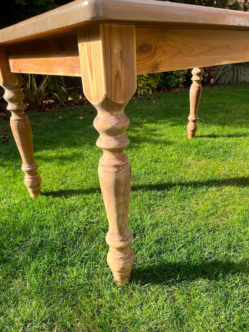 May include: A light brown wooden table with a rectangular top and four ornate legs. The table is set on a green grassy surface. The legs have a turned design, adding a touch of elegance to the piece.
