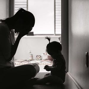 Pode incluir: Uma silhueta de uma pessoa sentada com a cabe&ccedil;a nas m&atilde;os, enquanto uma crian&ccedil;a senta-se no ch&atilde;o &agrave; sua frente. A imagem est&aacute; em preto e branco.